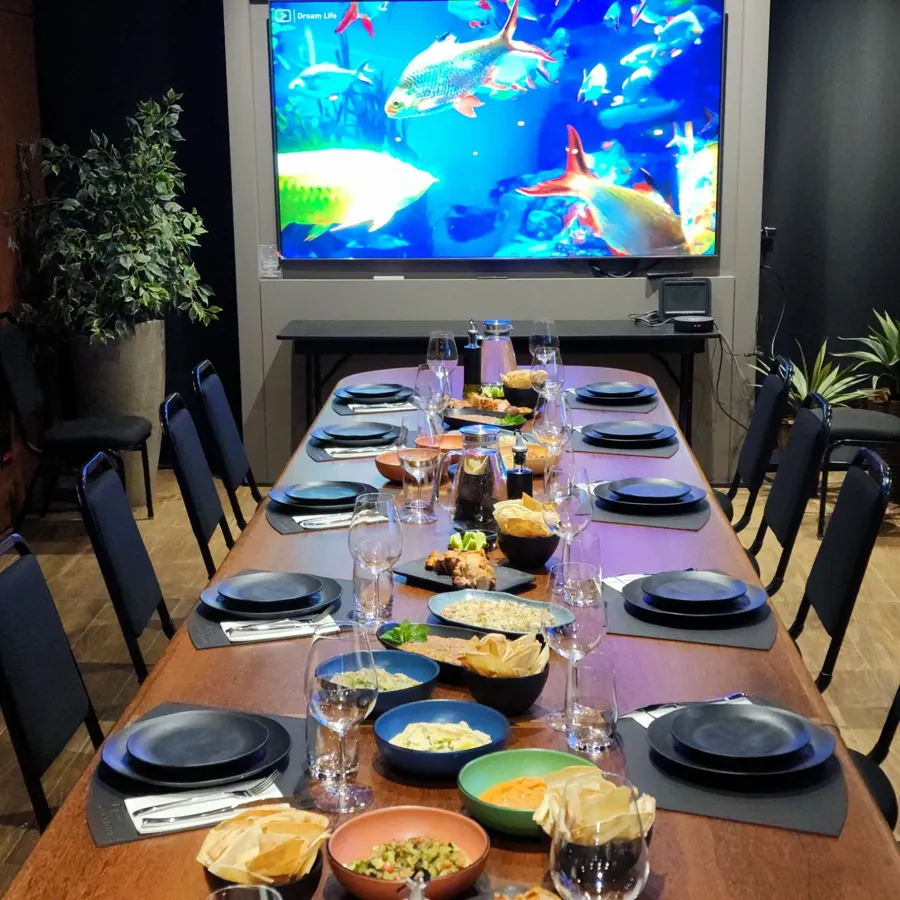 Mesa longa posta com pratos árabes na sala do Espaço Convivium, com TV de 100 polegadas ao fundo